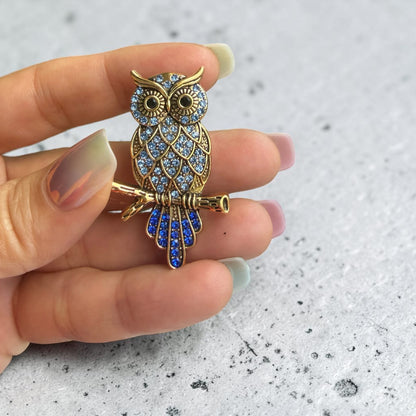 A gold owl brooch embellished with light blue and dark blue rhinestones, held on a person's fingers against a light gray textured background.