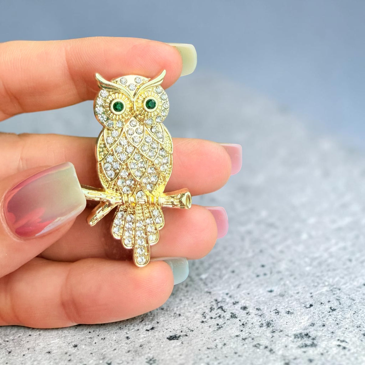 A gold owl brooch covered in clear rhinestones with green eye accents, held on a person's fingers over a gray textured surface.