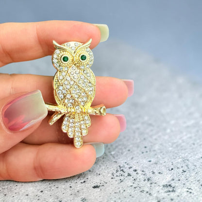 A gold owl brooch covered in clear rhinestones with green eye accents, held on a person's fingers over a gray textured surface.