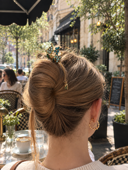 A woman viewed from behind at an outdoor cafe, wearing a gold hair accessory with blue-green floral details and a dangling charm in her hair bun.