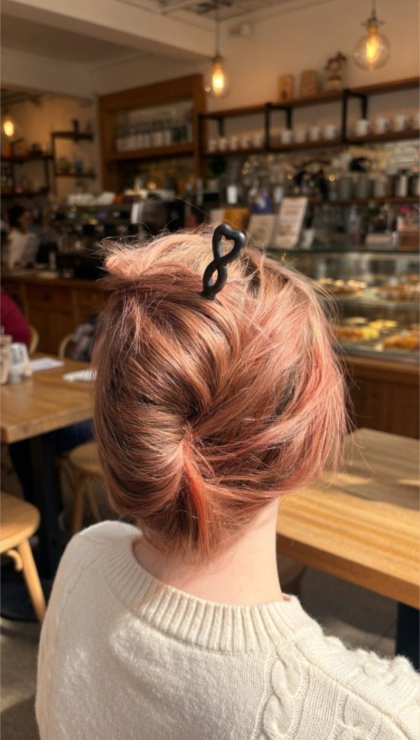 A person seated in a cafe, shown from behind, wearing a cream cable-knit sweater and a black hair clip holding up a messy bun hairstyle with peachy pink and dark brown hair.