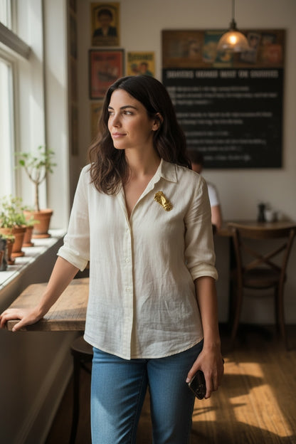 Luna Cotton abstract gold-tone face brooch pinned to a woman wearing a white linen shirt in a sunlit cafe setting.
