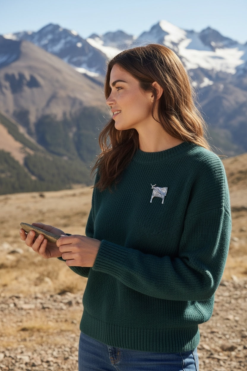 Luna Cotton silver Highland cow lapel pin worn on a woman's dark green knit jumper against a mountain landscape.