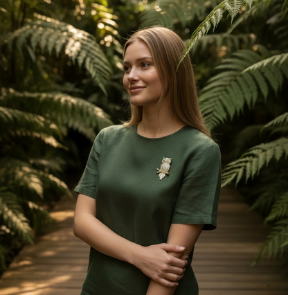 A woman wearing a dark green short-sleeve t-shirt with a small owl brooch on the chest, standing on a wooden boardwalk surrounded by lush green ferns.